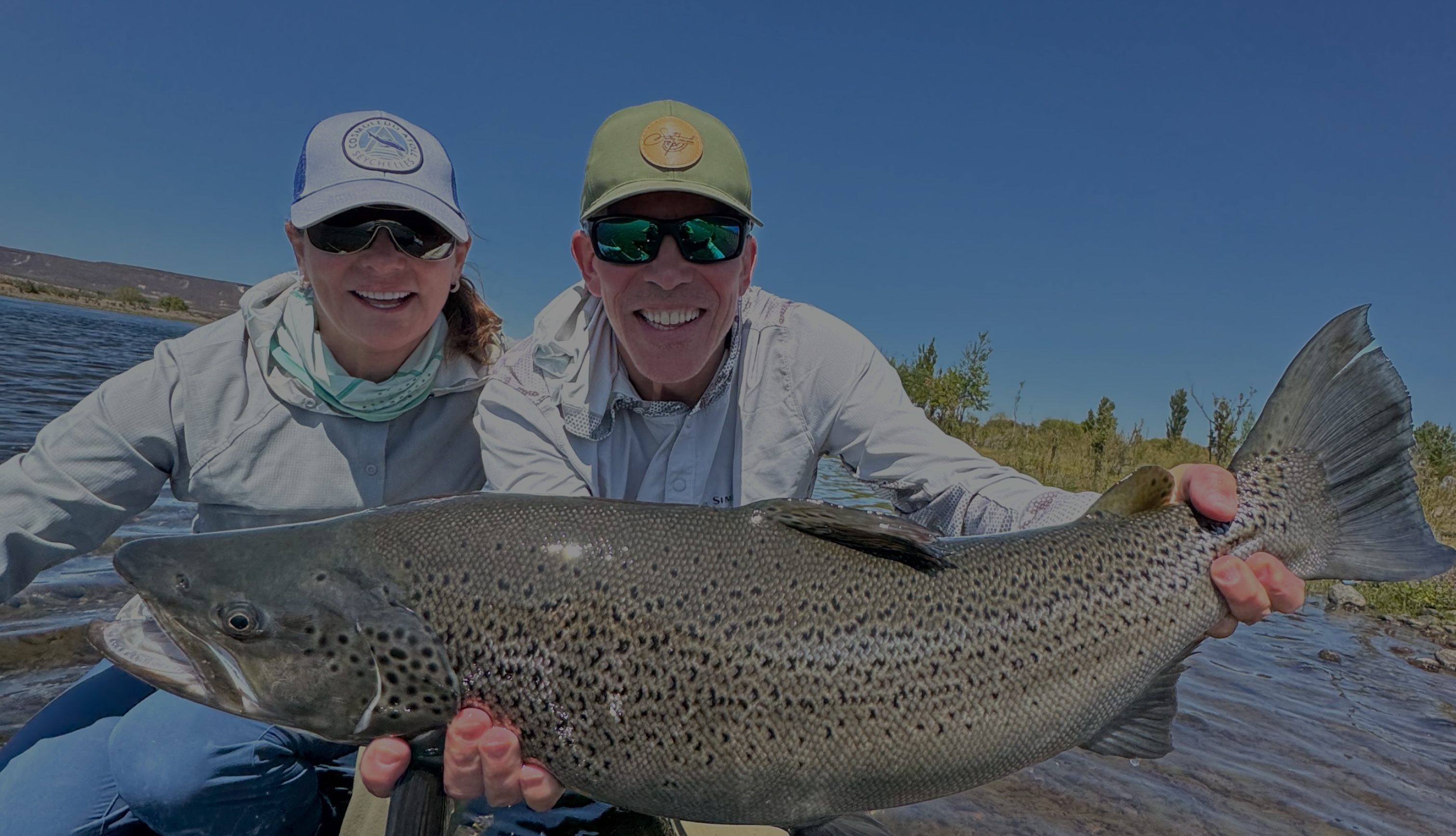 Two anglers holding a large trout at the water&rsquo;s edge